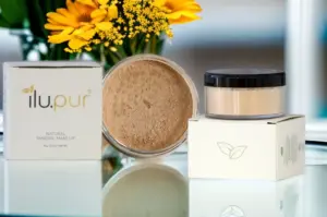 hautquartier Ein geöffneter Tiegel mit Natural Mineral Make-Up Warmton Nr. 4 – ilu.pur steht neben einem geschlossenen Tiegel und Schachteln, umgeben von gelben Blumen und verschwommenen grünen Blättern, die für eine frische Atmosphäre sorgen; das Ganze steht auf einer reflektierenden Oberfläche.